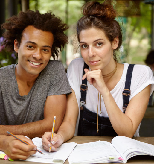 Handsome black male student and attractive female with European appearance working together writing in textbooks searching information for diploma paper while sitting at wooden desk of cozy cafe Handsome black male student and attractive female with European appearance working together writing in textbooks searching information for diploma paper while sitting at wooden desk of cozy cafe